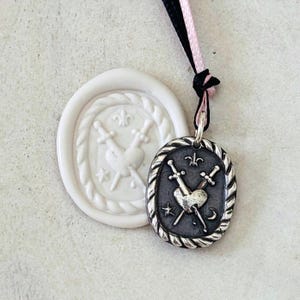 May include: A silver pendant with an oval shape, featuring a heart pierced by two swords, a fleur-de-lis, a crescent moon, and stars. The pendant hangs from a black and pink cord. Beside it is a white wax seal with a similar design.