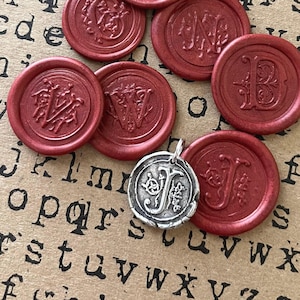 May include: A collection of red wax seals with various initial designs and a silver pendant with the letter "J". The seals are round and embossed with ornate letters. The pendant is also round and features the letter "J" with decorative elements.