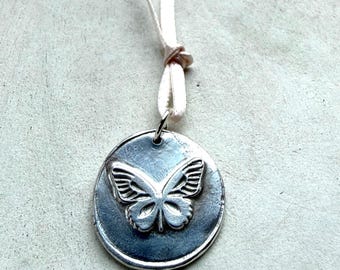 May include: A circular silver-toned metal pendant featuring a raised butterfly design in the center. The pendant has a dark, oxidized finish in the recessed areas and is suspended from a thin, light-colored ribbon tied in a simple knot at the top.