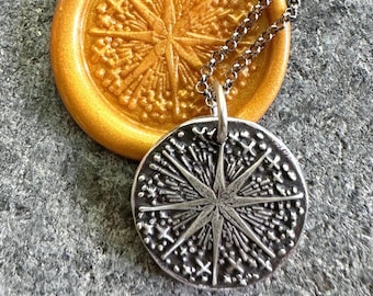May include: A round silver-toned pendant featuring a raised starburst design with radiating lines and small cross shapes, suspended from a silver-toned chain. The pendant rests against a circular gold-toned wax seal with a matching starburst pattern.