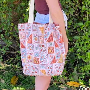 以下が含まれることがあります： 女性がカラフルな幾何学模様の白いキャンバス地のトートバッグを持っている。バッグは茶色のストラップで、赤、オレンジ、黄色、青、緑の幾何学模様で飾られている。