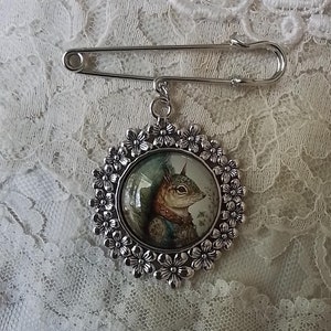 May include: A silver brooch with a round, floral-patterned frame. Inside the frame is a circular image of a squirrel wearing a blue and white patterned vest.