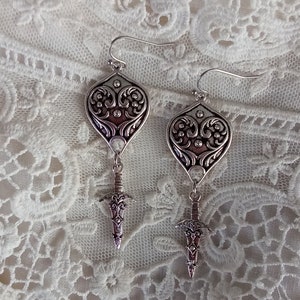 May include: A pair of silver earrings with a heart-shaped design and a sword pendant. The earrings are on a white lace background.