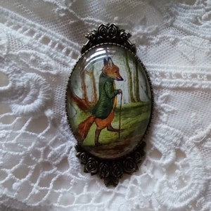 May include: An oval-shaped brooch with a vintage aesthetic. The brooch features a detailed illustration of a fox in a green coat and holding a cane, set against a forest backdrop. The brooch is framed by an ornate, dark metal border, and rests on a white lace surface.