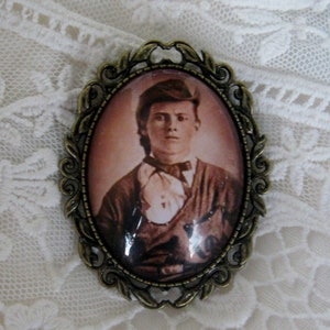 May include: A vintage-style bronze-toned brooch with a glass cameo featuring a portrait of a man wearing a bow tie and a dark jacket.