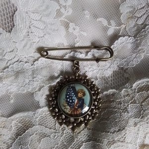 May include: Vintage-style brooch with a patriotic theme. The brooch features a circular pendant with a child holding an American flag. The pendant is surrounded by a floral design and attached to a safety pin clasp.