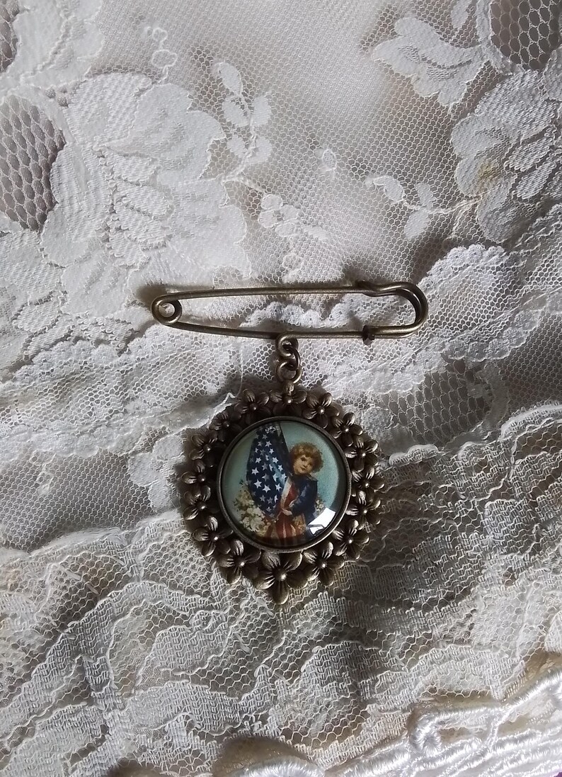 May include: Vintage-style brooch with a patriotic theme. The brooch features a circular pendant with a child holding an American flag. The pendant is surrounded by a floral design and attached to a safety pin clasp.