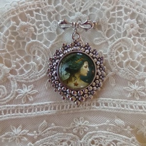 Pode incluir: Um broche de prata com um design floral e um camafeu redondo de vidro que apresenta uma mulher com cabelo comprido e escuro e uma coroa. O camafeu est&aacute; engastado em uma moldura de prata com um design floral.