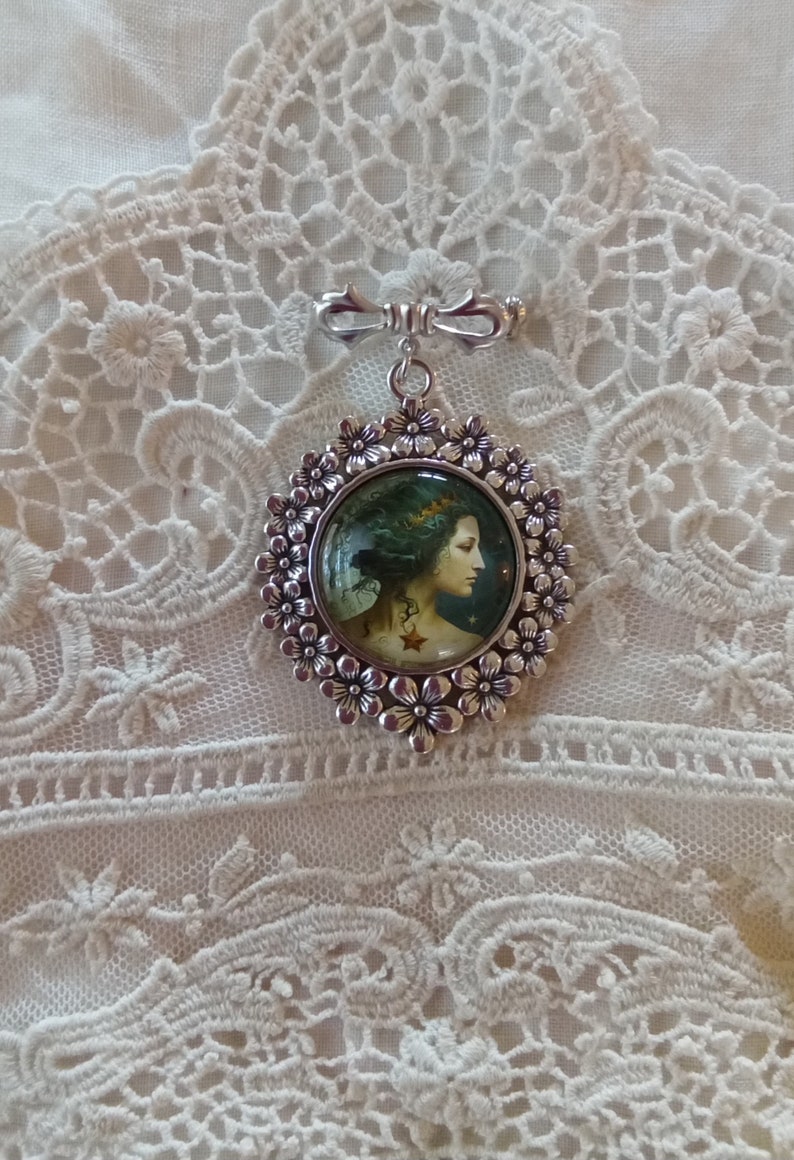 Pode incluir: Um broche de prata com um design floral e um camafeu redondo de vidro que apresenta uma mulher com cabelo comprido e escuro e uma coroa. O camafeu est&aacute; engastado em uma moldura de prata com um design floral.