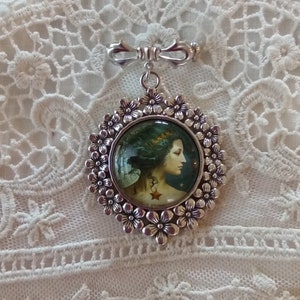 Pode incluir: Um broche de prata com um camafeu redondo de vidro que apresenta uma mulher com cabelo comprido e escuro e uma coroa de folhas. O camafeu &eacute; rodeado por uma moldura de prata com um design floral.