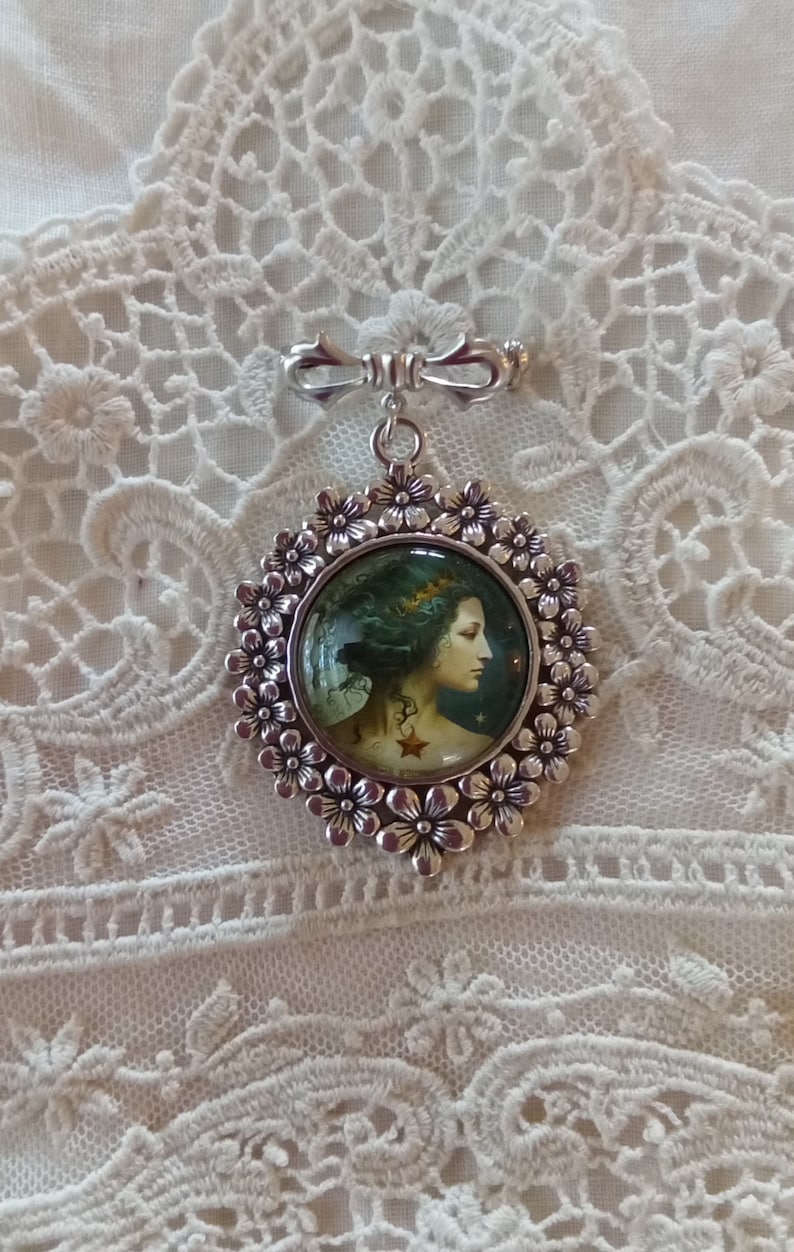 Pode incluir: Um broche de prata com um camafeu redondo de vidro que apresenta uma mulher com cabelo comprido e escuro e uma coroa de folhas. O camafeu &eacute; rodeado por uma moldura de prata com um design floral.