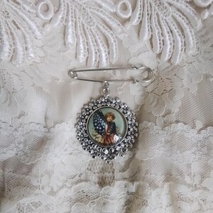 May include: A silver-toned brooch with a floral border and a patriotic image of a child holding an American flag. The brooch is attached to a safety pin, set against a white lace background.