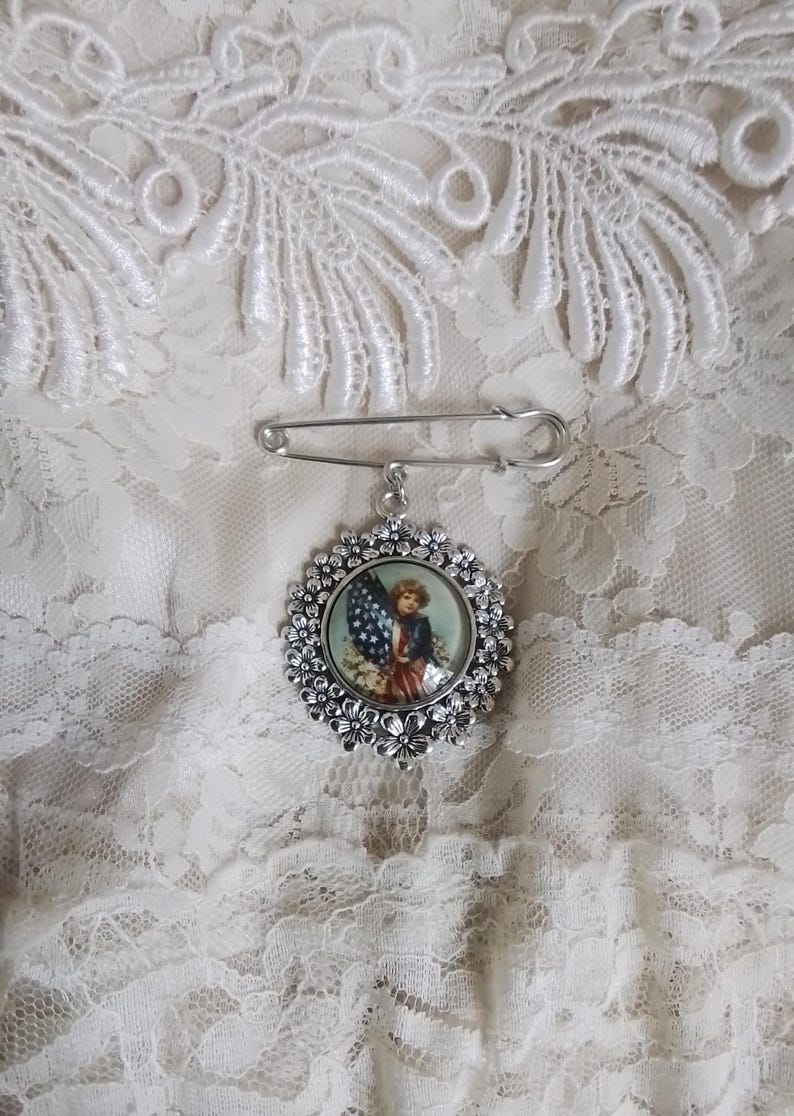 May include: A silver-toned brooch with a floral border and a patriotic image of a child holding an American flag. The brooch is attached to a safety pin, set against a white lace background.