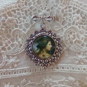 Pode incluir: Um broche de prata com um camafeu redondo que apresenta uma mulher com cabelo escuro e uma coroa. O camafeu &eacute; rodeado por um c&iacute;rculo de flores de prata.