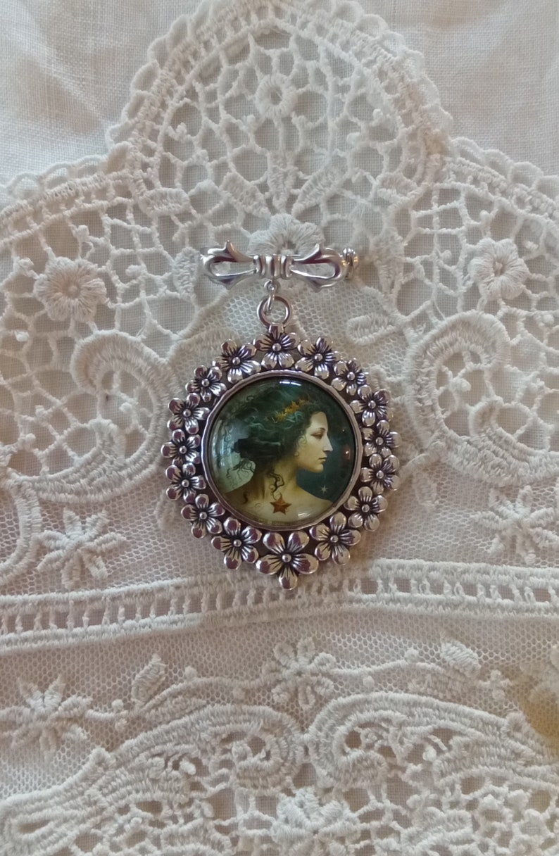 Pode incluir: Um broche de prata com um camafeu redondo que apresenta uma mulher com cabelo escuro e uma coroa. O camafeu &eacute; rodeado por um c&iacute;rculo de flores de prata.