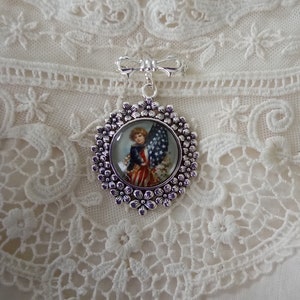 May include: A silver-toned brooch featuring a patriotic design. The brooch has a circular pendant with a vintage illustration of a child holding an American flag. The pendant is surrounded by a floral border and a bow-shaped top.