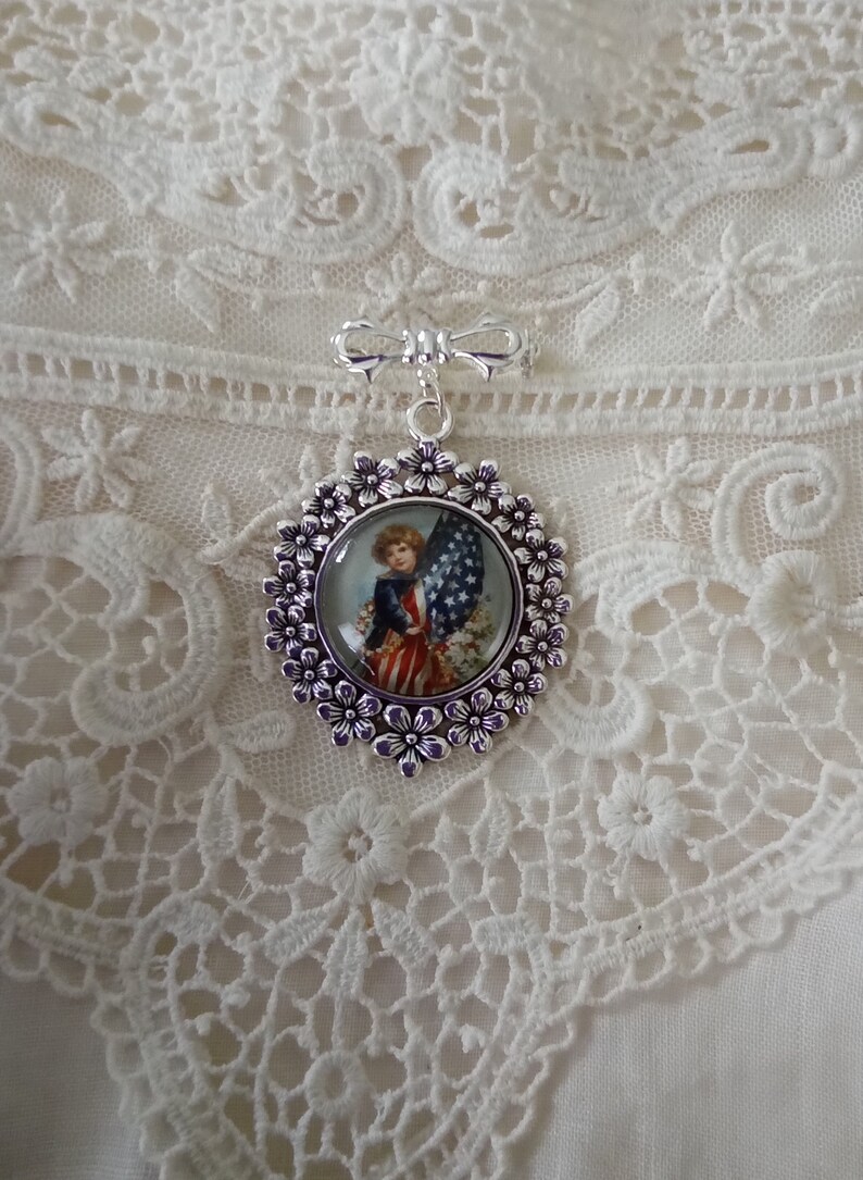 May include: A silver-toned brooch featuring a patriotic design. The brooch has a circular pendant with a vintage illustration of a child holding an American flag. The pendant is surrounded by a floral border and a bow-shaped top.