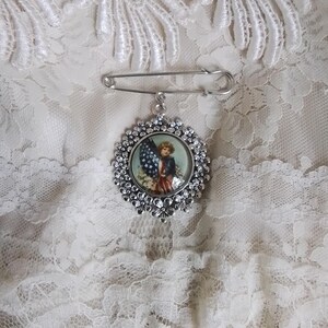 May include: A silver-toned brooch with a floral border and a patriotic image of a child holding an American flag. The brooch is attached to a large safety pin, set against a white lace background.
