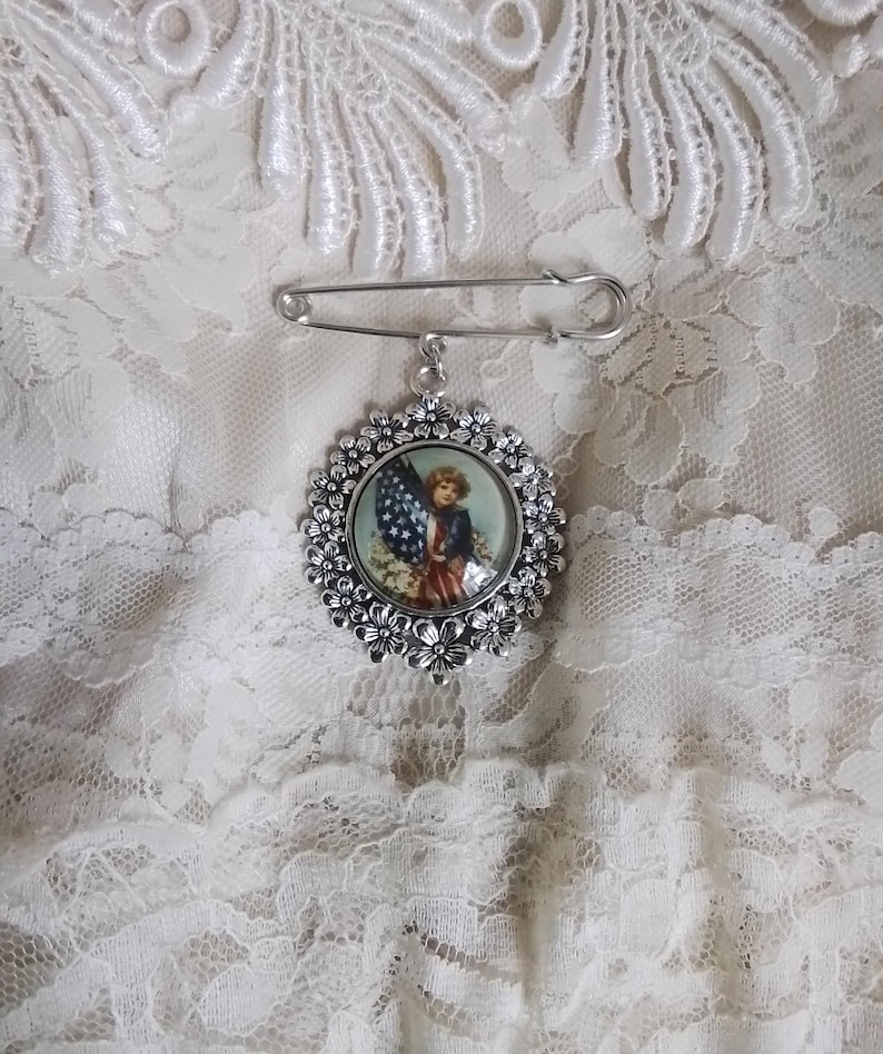 May include: A silver-toned brooch with a floral border and a patriotic image of a child holding an American flag. The brooch is attached to a large safety pin, set against a white lace background.