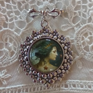Pode incluir: Um broche de prata com um camafeu redondo que apresenta uma mulher com cabelo comprido e escuro e uma estrela no ombro. O camafeu &eacute; rodeado por uma moldura de prata com um design floral.
