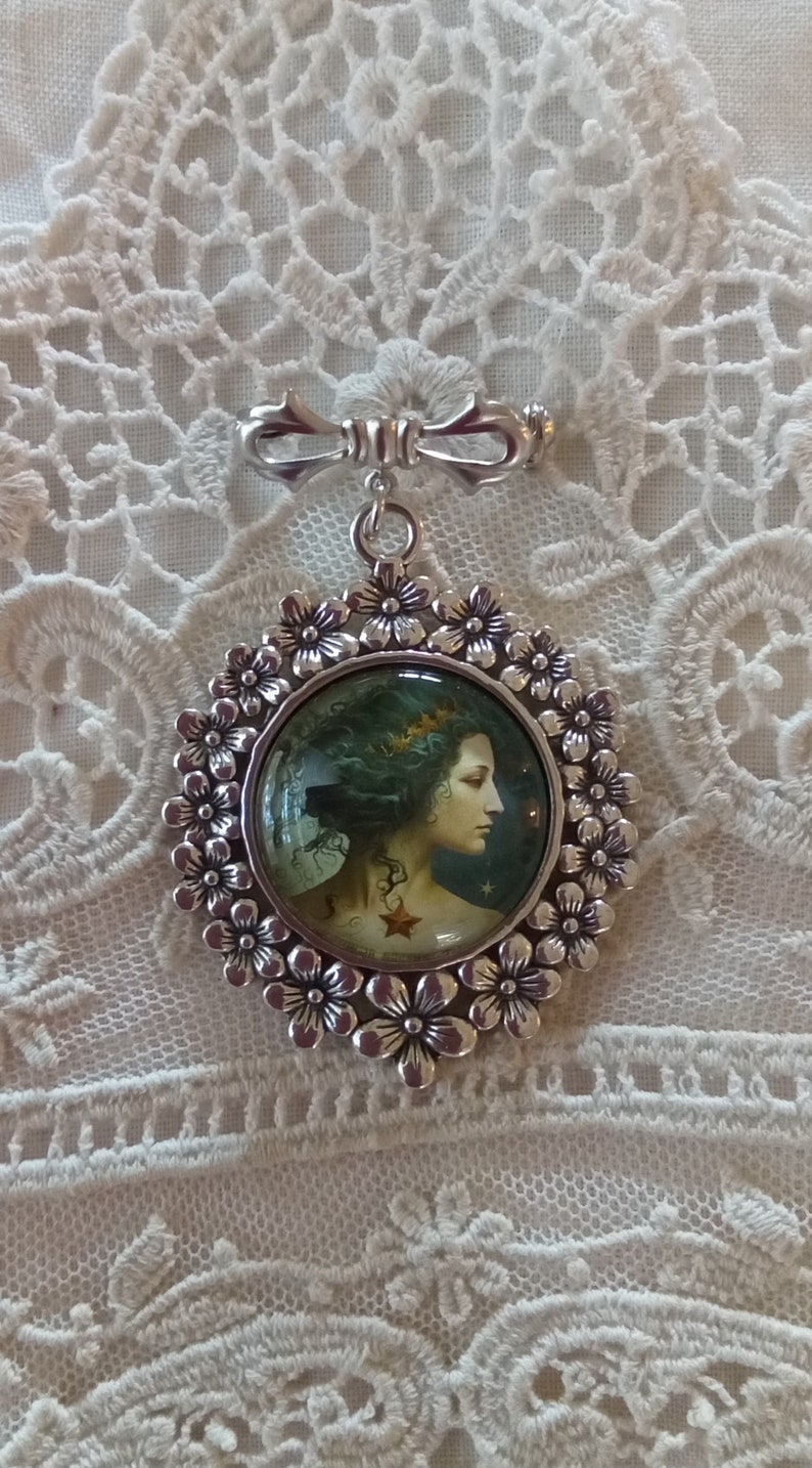 Pode incluir: Um broche de prata com um camafeu redondo que apresenta uma mulher com cabelo comprido e escuro e uma estrela no ombro. O camafeu &eacute; rodeado por uma moldura de prata com um design floral.