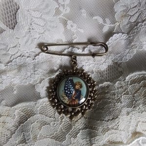 May include: Vintage patriotic brooch featuring a child holding an American flag. The round pendant has a floral border and is attached to a safety pin clasp. The image inside the pendant has red, white, and blue colors.