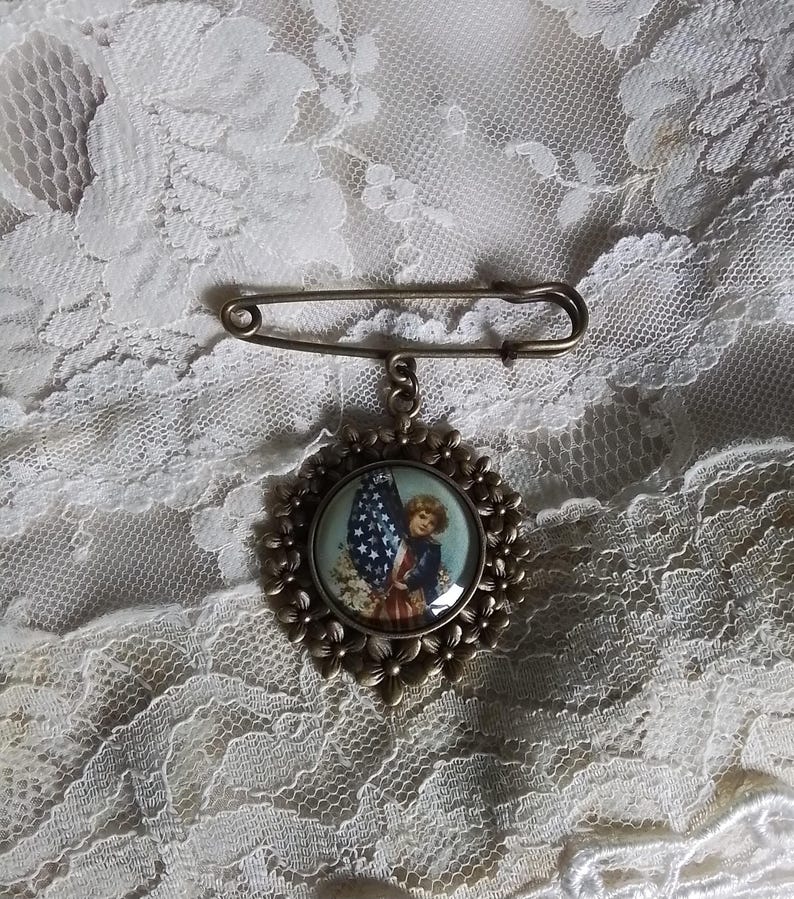 May include: Vintage patriotic brooch featuring a child holding an American flag. The round pendant has a floral border and is attached to a safety pin clasp. The image inside the pendant has red, white, and blue colors.