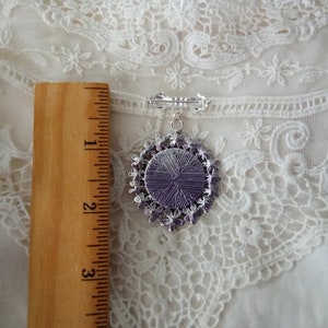 May include: A silver-toned brooch with a pendant featuring a purple center and a decorative, ornate border. The brooch is approximately 2 inches long, with a pin on the back for secure attachment. The background is a white, lacy fabric.