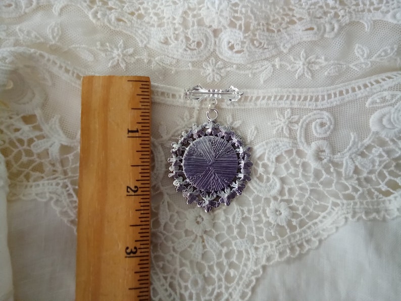 May include: A silver-toned brooch with a pendant featuring a purple center and a decorative, ornate border. The brooch is approximately 2 inches long, with a pin on the back for secure attachment. The background is a white, lacy fabric.