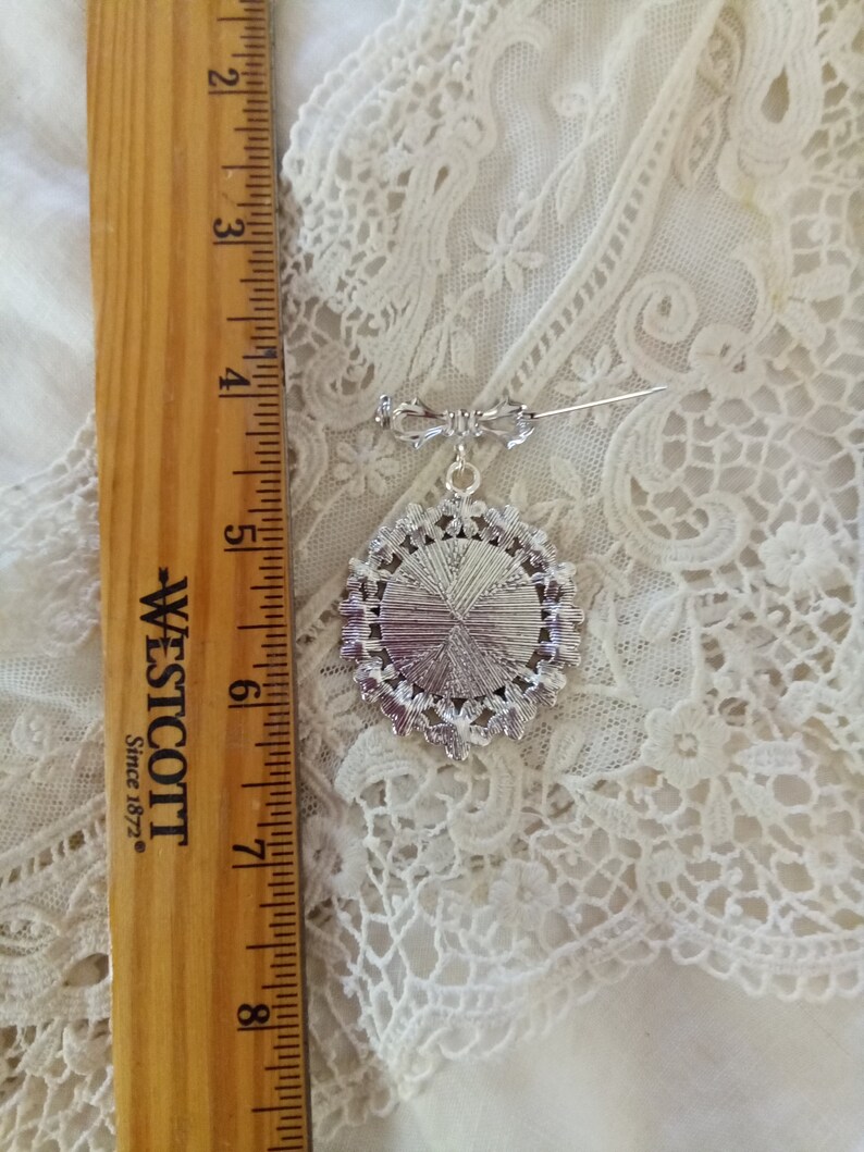 Pode incluir: Um broche de prata com um design redondo e ornamentado. O broche tem aproximadamente 5 cm de di&acirc;metro e tem um pino na parte de tr&aacute;s.