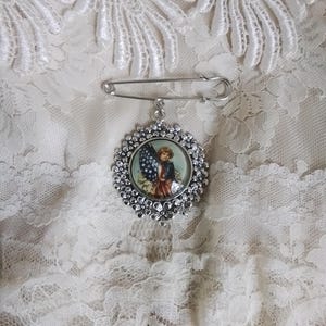 May include: A silver-toned brooch with a decorative floral border and a pin closure. The brooch features a circular image of a child holding an American flag. The brooch is set against a white lace background.