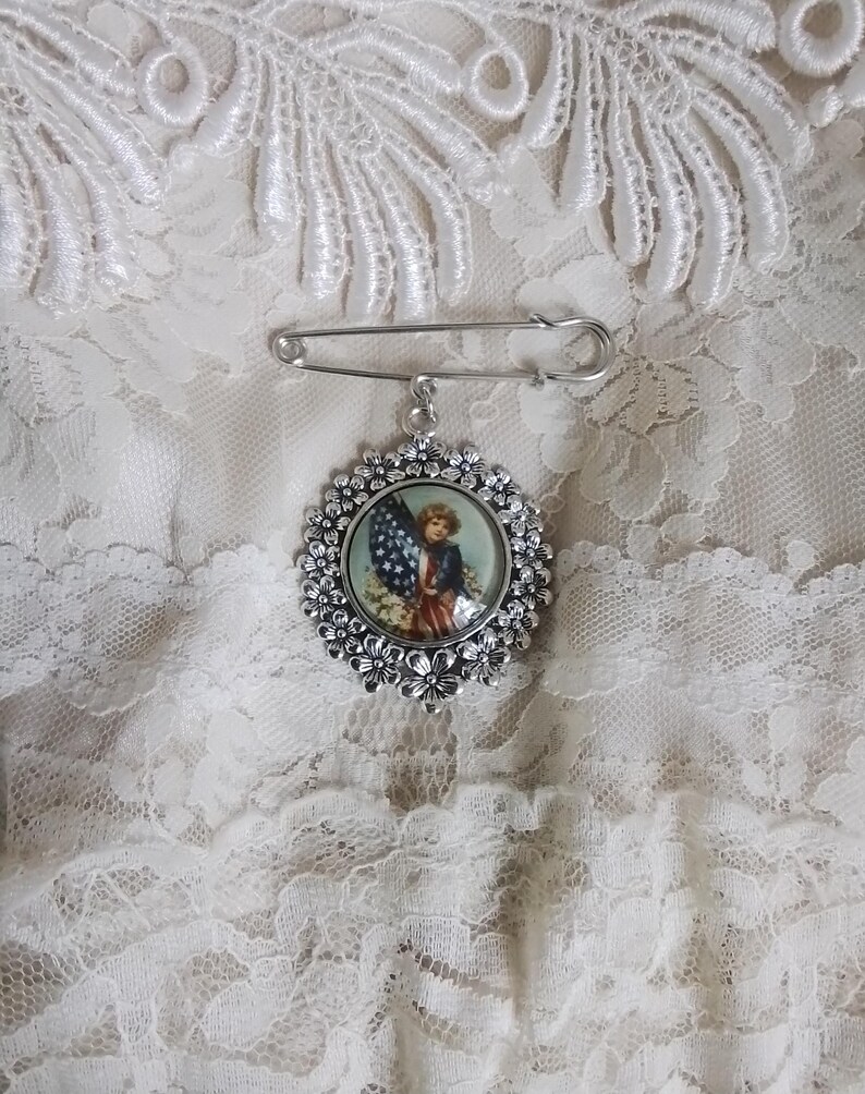 May include: A silver-toned brooch with a decorative floral border and a pin closure. The brooch features a circular image of a child holding an American flag. The brooch is set against a white lace background.