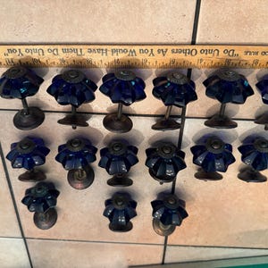 May include: Fourteen cobalt blue glass knobs with a faceted, flower-like shape and dark metal bases. A wooden ruler is positioned above the knobs, showing a scale of 12 inches. The knobs are arranged in three rows on a light-colored tiled surface.