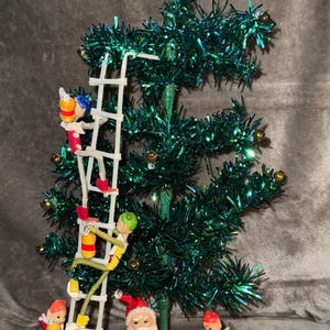 May include: A miniature Christmas tree with green tinsel branches, adorned with small gold ornaments. The tree is decorated with figurines of Santa Claus, elves, and an angel at the top. The elves are climbing ladders.