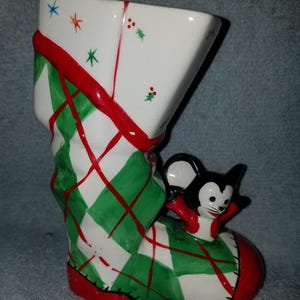 May include: A ceramic Christmas stocking with a green and red plaid pattern and a small mouse figurine on the side. The stocking is white with red trim and has a red base.
