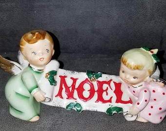 May include: Two porcelain angel figurines holding a banner that reads "NOEL" in red letters. The angels are dressed in green and pink with gold wings and holly accents.