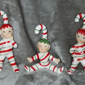 May include: Three ceramic Christmas elf figurines with candy cane hats. The elves are wearing red and white striped outfits with green accents.