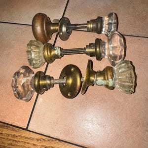 May include: Three vintage door knobs with brass hardware and clear glass handles. The set includes one round wooden knob, one fluted glass knob, and one faceted glass knob, each featuring a metal spindle and circular mounting plates with screw holes.
