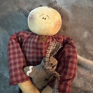 May include: A handmade doll with a round, cream-colored head and wire hair. The doll wears a burgundy plaid shirt and holds a gray gingerbread man with a plaid scarf. The doll's face has simple stitched features.