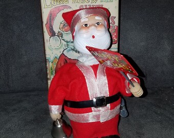 May include: A red and white Santa Claus figure with a white beard and a red hat. He is holding a sign that says "Merry Christmas" and a bell. The figure is a wind-up toy and is part of a set called "Long-Neck the Santa".
