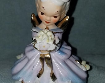 May include: A porcelain figurine of a little angel girl with blonde hair, wearing a purple and blue dress with gold trim. She is holding a bouquet of white flowers and has a gold halo. The base of the figurine is white with the words "Monday's Child" written in gold.