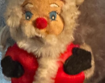 May include: A vintage Santa Claus doll with a red suit, black gloves, and a red hat. The doll has a tan face with blue eyes, a red nose, and a white beard and trim. The doll is made of fabric and has a soft, plush texture.