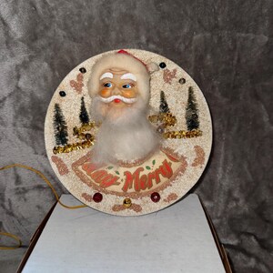 May include: Vintage Christmas decoration featuring Santa Claus with a white beard, blue eyes, and a red hat. The circular ornament has a glittery background with the words "Day Merry" and decorative trees.