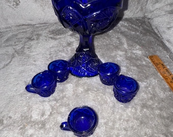 Puede incluir: Un cuenco de pedestal de vidrio azul cobalto con un borde festoneado y patrones geométricos prensados, acompañado de cinco tazas en miniatura con asa a juego. Una regla de madera es visible a la derecha, mostrando medidas en centímetros.