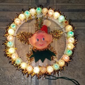 May include: A vintage Christmas tree topper featuring a smiling elf with a green hat and a gold tinsel garland with multi-colored lights.
