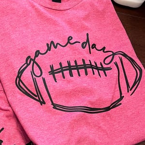 May include: A pink t-shirt featuring the words "game day" in a cursive font and a black football graphic. The shirt is made of a soft material, ideal for game day.