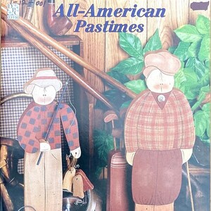 Puede incluir: Figuras de arte popular de madera que representan actividades de ocio. Una figura sostiene una caña de pescar, la otra un palo de golf. Las figuras visten camisas y sombreros a cuadros. La imagen se titula "All-American Pastimes."