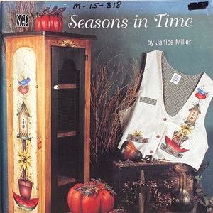 May include: A wooden cabinet with painted folk art, a white vest with painted designs, and several pumpkins. The cabinet has a black mesh door and is decorated with a bird, heart, and birdhouse. The vest features a birdhouse, sunflower, and anchor design. The pumpkins are red.