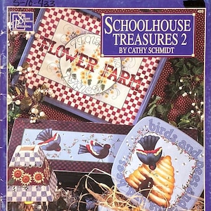 May include: A collection of decorative items with a country theme. The image features a book titled "Schoolhouse Treasures 2" and several painted decorative pieces, including a lamp, a plate, and a wooden box. The plate has the text "birds and bees and spring".