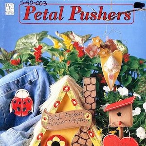 May include: A colorful wooden birdhouse with a red roof and a yellow flower painted on the door. The birdhouse has a sign that says "Petal Pusher's Flower Shoppe". There is a red heart sign that says "Open" hanging from a birdhouse on a stick. The birdhouse is surrounded by green plants and flowers.